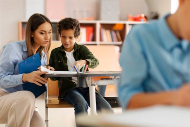 Young female teacher assisting Latin schoolboy, helping with task in test. Education, learning, childhood, teaching and school concept