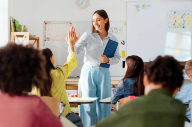 Happy friendly female teacher sharing high-five with girl, fostering positive learning environment in classroom of private school