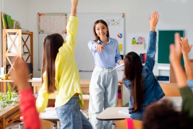 Cheerful female teacher teaching secondary school pupils, kids raising hands while teacher asks questions, classroom interior