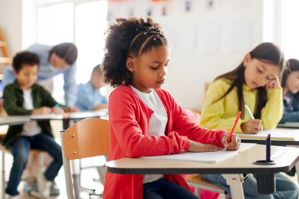African American schoolgirl writing in copybook in classroom at elementary school, children sitting at desks, learning at lesson subject with teacher