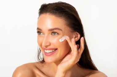 Woman putting cream on fresh soft pure skin and smiling, lady with natural makeup applying natural beauty cosmetics product over white background