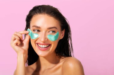 Authentic European woman with eye patches looking away on copy space, standing isolated on white background. Skin care, cosmetic product concept