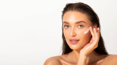 Beautiful lady with cream on face, touching cheek with fingers, posing over white studio background. Skincare and cosmetology concept
