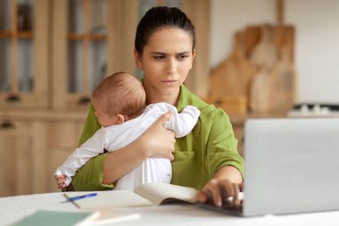 Yorgun anne kollarında kaprisli bebekle çalışmaya çalışır dizüstü bilgisayar klavyesinde yazı yazar, mutfakta masada oturur.