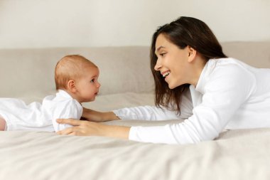 Mutlu anne küçük kızıyla oynuyor, karnının üstünde yatıyor, sabahını yatak odasında geçiriyor, yan görüş açısıyla. Çocuk bakımı, mutlu annelik.