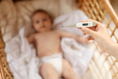 Termometreyi tutan şefkatli bir anne, beşikte yatan hasta bir bebeğin sıcaklığını ölçüyor. Çocuk hastalıkları, çocuk bakımı konsepti