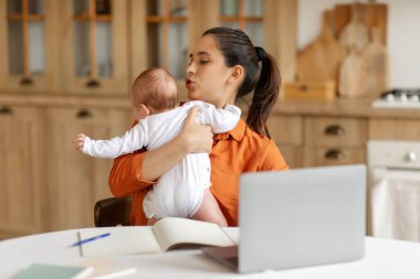 Evde bebek sahibi bir iş kadını dizüstü bilgisayar kullanıyor, kucağında ağlayan bebekle masada oturuyor, boşluğu kopyalıyor.