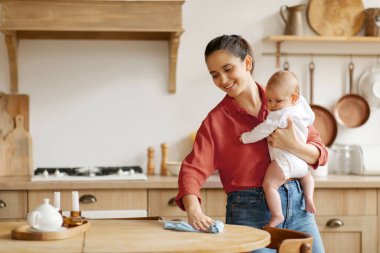 Ebeveynlik ve çoklu görev. Anne kucağında küçük bir bebek, mutfakta temizlik masası, ev işleri, boş yer.