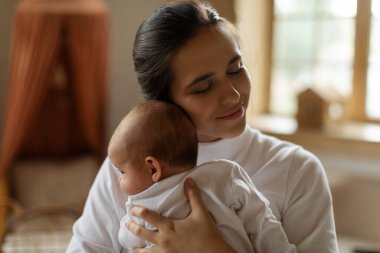Anne ve bebek arasında bir bağ. Anne küçük bebeğini nazikçe kucaklıyor, evdeki rahat yatak odasında kaynaşıyor.