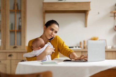 Cep telefonuyla iletişim kuran serbest çalışan bir kadın dizüstü bilgisayarla çalışıyor ve evde bebeğine bakıyor, mutfakta masada oturuyor.