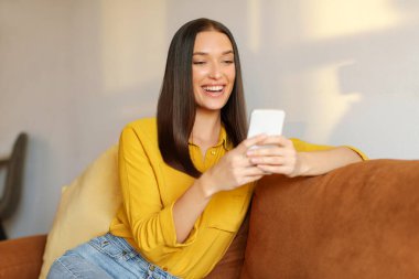 Mutlu genç bir kadın rahat koltukta dinleniyor, elinde akıllı telefon, sosyal ağlarda sohbet ediyor, video izliyor ya da mobil uygulamalar kullanıyor.