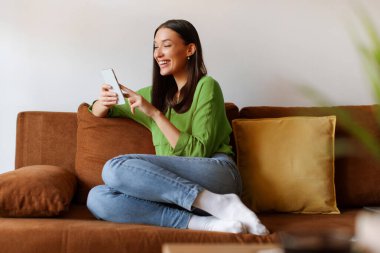 Pozitif kadın koltukta oturuyor, cep telefonu kullanıyor, gülümsüyor, ekrana bakıyor, online ekommerce alışverişi yapıyor ya da video seyrediyor.