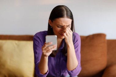 Akıllı telefonu tutan üzgün bir kadın, üzgün, stresli bir kadın. Hoş olmayan bir mesaj aldı. Evdeki kanepede oturuyor.