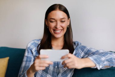 Cep telefonuyla oyun oynayan heyecanlı bir kadın, evdeki rahat koltukta eğlenen, mutlu ve rahat hisseden bir kadın.