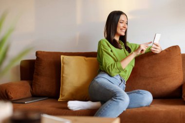 Evdeki kanepede dinlenirken akıllı telefon kullanan gülümseyen bayan, online alışveriş için cep telefonu kullanıyor, sosyal ağlara bakıyor ya da mesajlaşıyor.