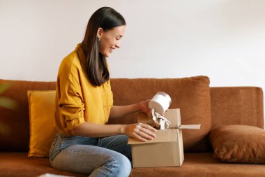 Karton güzellik kutusunu kozmetik malzemelerle açıp gülümseyen, evdeki oturma odasında kanepede oturan tatmin olmuş genç bir kadın.