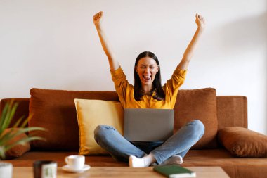Heyecanlı serbest çalışan kadın dizüstü bilgisayarla koltukta oturuyor, ellerini kaldırıyor, EVET işareti yapıyor ve başarıyı kutluyor, boş alan. Alışveriş satışı, terfi, başarı konsepti
