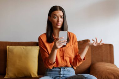 Kafası karışmış bir kadın evde koltukta otururken akıllı telefona bakıyor cep telefonundan mesajlar okuyor, haberlere duygusal tepki veriyor.