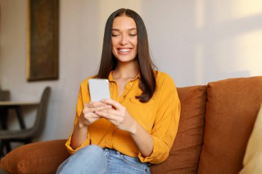 Gülümseyen Avrupalı kadın cep telefonunu iletişim için kullanıyor, evdeki kanepede dinleniyor, mesajlaşıyor ya da internette geziniyor.