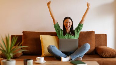 Heyecanlı serbest çalışan kadın dizüstü bilgisayarla koltukta oturuyor, ellerini kaldırıyor, EVET işareti yapıyor ve başarıyı kutluyor, boş alan. Alışveriş satışı, terfi, başarı konsepti