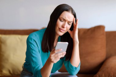 Endişeli kadın elinde akıllı telefon, dokunaklı kafa, evdeki koltukta oturan hayal kırıklığına uğramış kadın hoş olmayan mesajlar okuyor, baş ağrısı çekiyor.