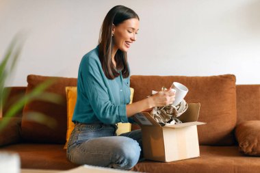 Memnuniyet dolu bayan paketini evde açıyor, neşeyle kutusunu açıyor ve kupayı keşfediyor, online alışveriş macerasının zevkini kucaklıyor.