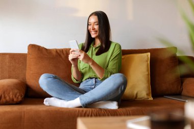 Kanepede otururken, internet alışverişi yaparken ya da arkadaşlarıyla mesajlaşırken akıllı telefonuna bakmaktan hoşlanan mutlu bir kadın.