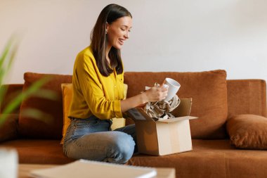 Memnuniyet dolu bayan paketini evde açıyor, neşeyle kutusunu açıyor ve kupayı keşfediyor, online alışveriş macerasının zevkini kucaklıyor.