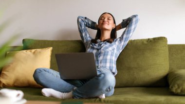 Kanepede oturan mutlu kadın ellerini başının arkasına koy rahat koltuğa uzan, dizüstü bilgisayar kullan, serbest çalışan işini bitir, rahatla ve hayal kur.