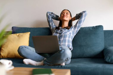 Kanepede oturan mutlu kadın ellerini başının arkasına koy rahat koltuğa uzan, dizüstü bilgisayar kullan, serbest çalışan işini bitir, rahatla ve hayal kur.