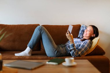 Ellerinde akıllı telefonuyla kanepeye uzanmış neşeli, rahat bir bayan, cep telefonu kullanırken gülümsüyor, arkadaşlarıyla mesajlaşıyor, rahatlık ve mutluluk yayıyor, bedava boşluk.