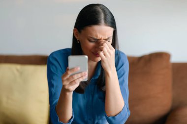 Akıllı telefonu tutan üzgün bir kadın, üzgün, stresli bir kadın. Hoş olmayan bir mesaj aldı. Evdeki kanepede oturuyor.