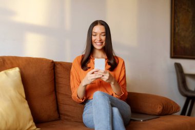 Akıllı telefondan mesajlaşan, evdeki koltukta oturan, sosyal ağlara ya da mesajlara bakan mutlu genç bir kadın.