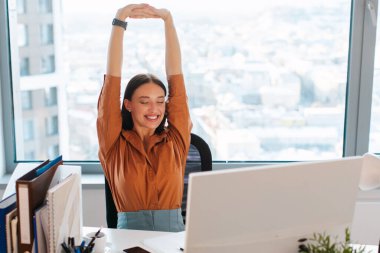 Heyecanlı genç iş kadını kollarını başının üstüne uzatıyor ve zevk içinde gülümsüyor, ofisinde masasında oturuyor.