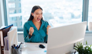 Heyecanlı bir profesyonel. Mutluluktan havalara uçan iş kadını iş zaferini kutluyor, bilgisayar monitörüne bakıyor, harika bir çalışmanın tadını çıkarıyor.