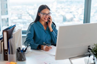 Mutlu kadın girişimci notlar alıyor ve müşteriyle cep telefonuyla konuşuyor, iş yerinde oturuyor ve iş planlarını tartışıyor.