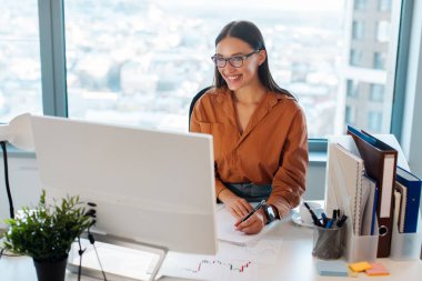 Çalışmak için masaüstü bilgisayarını kullanan mutlu bir kadın, sanal web sitesi ya da toplantısıyla görüntülü konuşma yapan, notlar yazan,