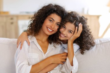 Sevimli küçük bir kız. Annesini kucaklıyor. Evdeki rahat koltukta oturuyor, anne ve kızını kucaklıyor, kameraya gülümsüyor.