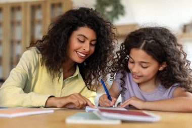 Sevgi dolu bir anne kızına okul ödevlerinde yardım ediyor, deftere yazıyor, evdeki rahat mutfak masalarında oturuyor.