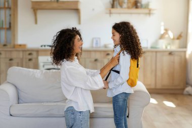 Latin anne kızını okul gününe hazırlıyor, çantaları omuzlara koymaya yardım ediyor, yandan bakıyor. Anne ve çocuk bağı, ebeveyn yardımı ve destek kavramı