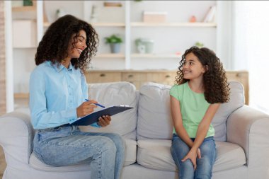 Mutlu çocuk kız psikolog ya da terapistle konuşuyor, kanepede oturuyor ve elinde dosya olan bir uzmanla konuşuyor. Çocuk psikolojisi ve terapi konsepti