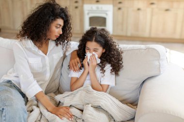 Hasta kızının yanında oturan şefkatli anne, burnunu silen kız, kendini kötü hisseden genç anne hasta kızına bakarken üzgün hissediyor.