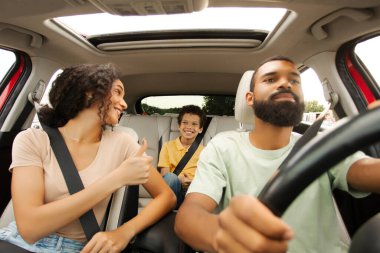 Mutlu siyahi ebeveynler ve oğlu araba yolculuğunun keyfini çıkarıyor, anne çocuğuna kemerini bağlayıp bakıyor, baş parmağını gösteriyor, baba araba sürüyor.