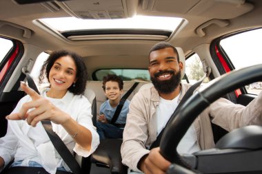 Neşeli Latin kadın aile gezisi sırasında bir şeyi işaret ediyor, erkek sürücü direksiyonda ve çocuk arka koltuktan izliyor, paylaşılan macerayı özetliyor.