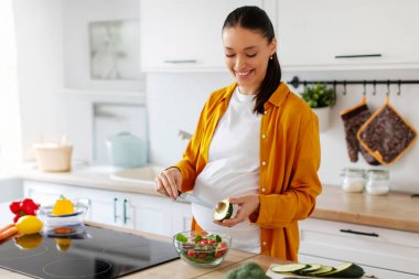 Genç hamile kadın sağlıklı yemek pişiriyor, salata için avokado soyuyor, ev mutfağında akşam yemeği hazırlıyor, boş yer var.