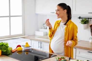 Mutfağın içinde sağlıklı yemek salatası hazırlarken taze sebze yiyen aşırı neşeli hamile kadın.