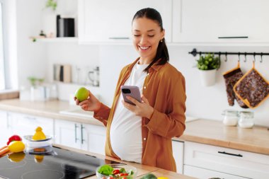 Akıllı telefonunu kullanan ve abur cubur yiyen mutlu, genç hamile bir kadın, iyi aydınlatılmış mutfakta elma yiyor.