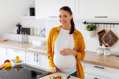 Neşeli, hamile bir kadın koca göbeğine dokunuyor ve kameraya gülümsüyor. Sağlıklı yemek hazırlarken mutfağın içinde duruyor. Sağlık, diyet, annelik kavramı