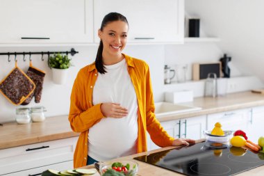 Karnına dokunan ve modern mutfağın içinde kameraya gülümseyen mutlu genç bir kadının portresi. Annelik, hamilelik süresince sağlıklı beslenme
