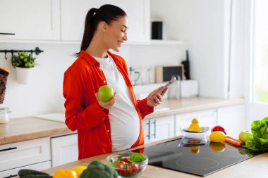 Mutlu Avrupalı bayan akıllı telefonuyla kitap okuyor ya da geziniyor ve tezgahta besleyici yiyeceklerle iyi aydınlatılmış mutfakta taze elma yiyor.